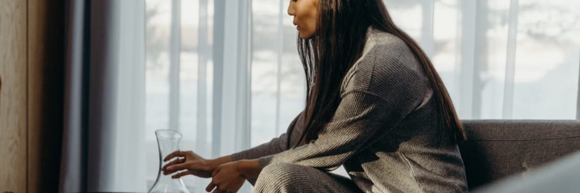 Woman sitting in a cozy room, pouring from a glass decanter on a sunny day.